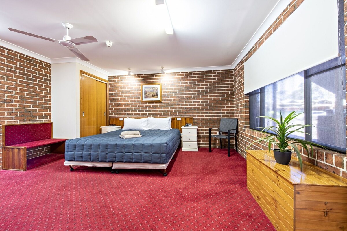 A bedroom with a brick wall, a bed, a desk, a chair, a wardrobe, and a window.