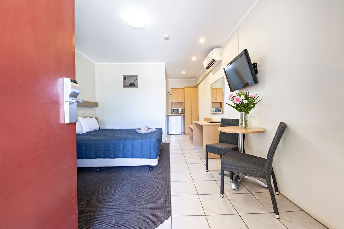 A hotel room with a bed, a desk, a chair, a TV, and a small kitchenette.