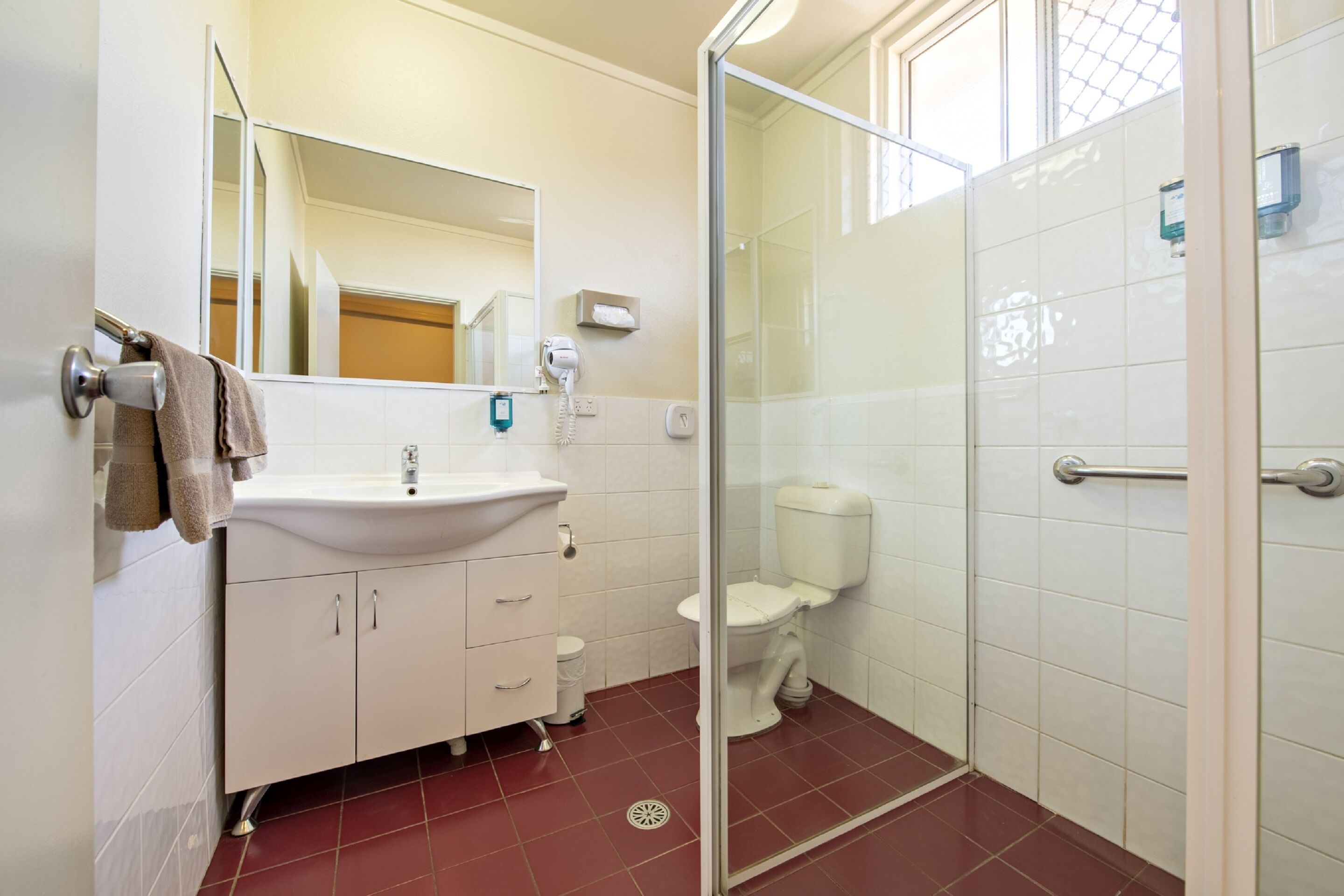 Un baño con lavamanos blanco, espejo, inodoro, puerta de ducha de cristal y suelo de baldosas rojas.