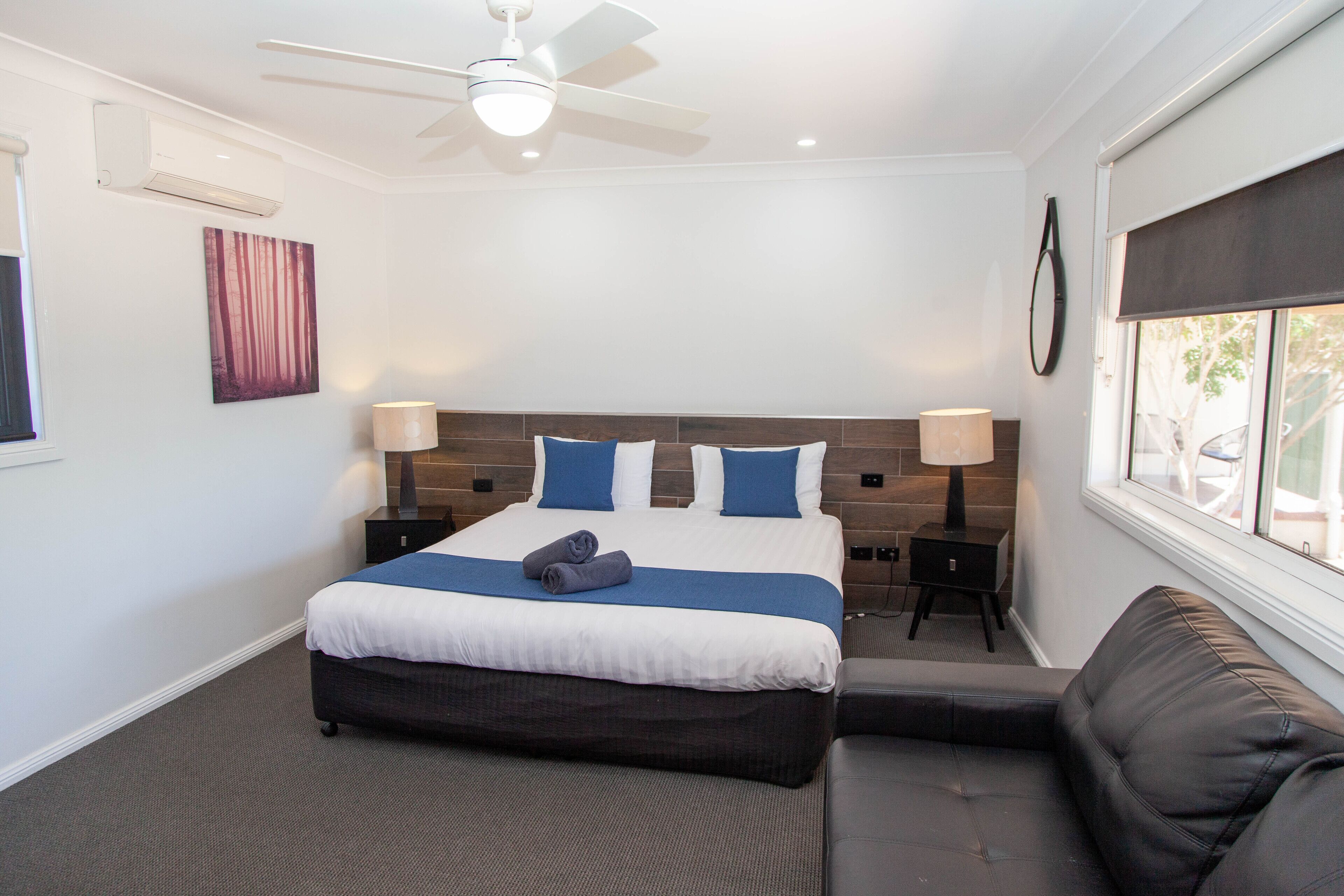 A modern hotel room with a large bed, a ceiling fan, a window with a view, and a small bedside table with a lamp.