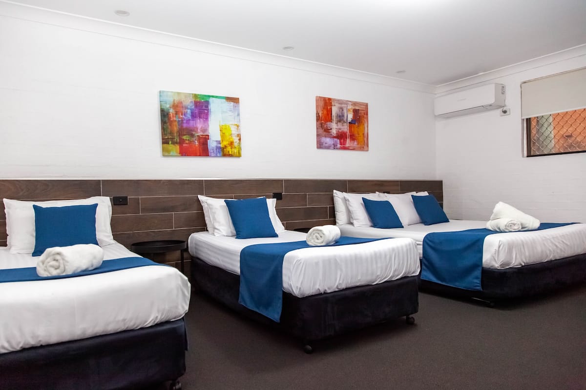 A hotel room with three beds, each with white linens and blue accents, a dark headboard, and abstract art on the walls.