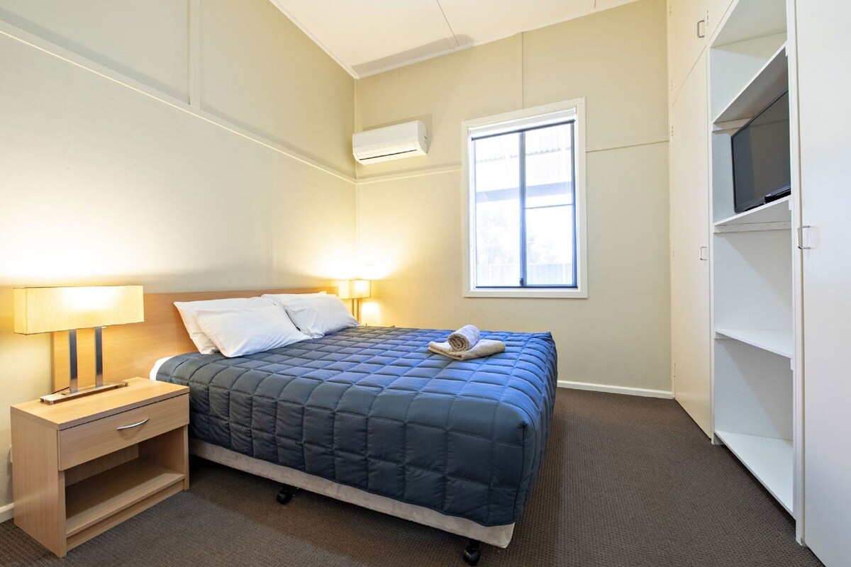 A bedroom with a bed, bedside table, lamp, air conditioning unit, window, and built-in wardrobe.