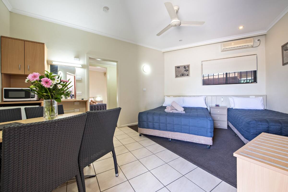 A hotel room with two beds, a dining area with a table and chairs, a microwave, a ceiling fan, and a large window.