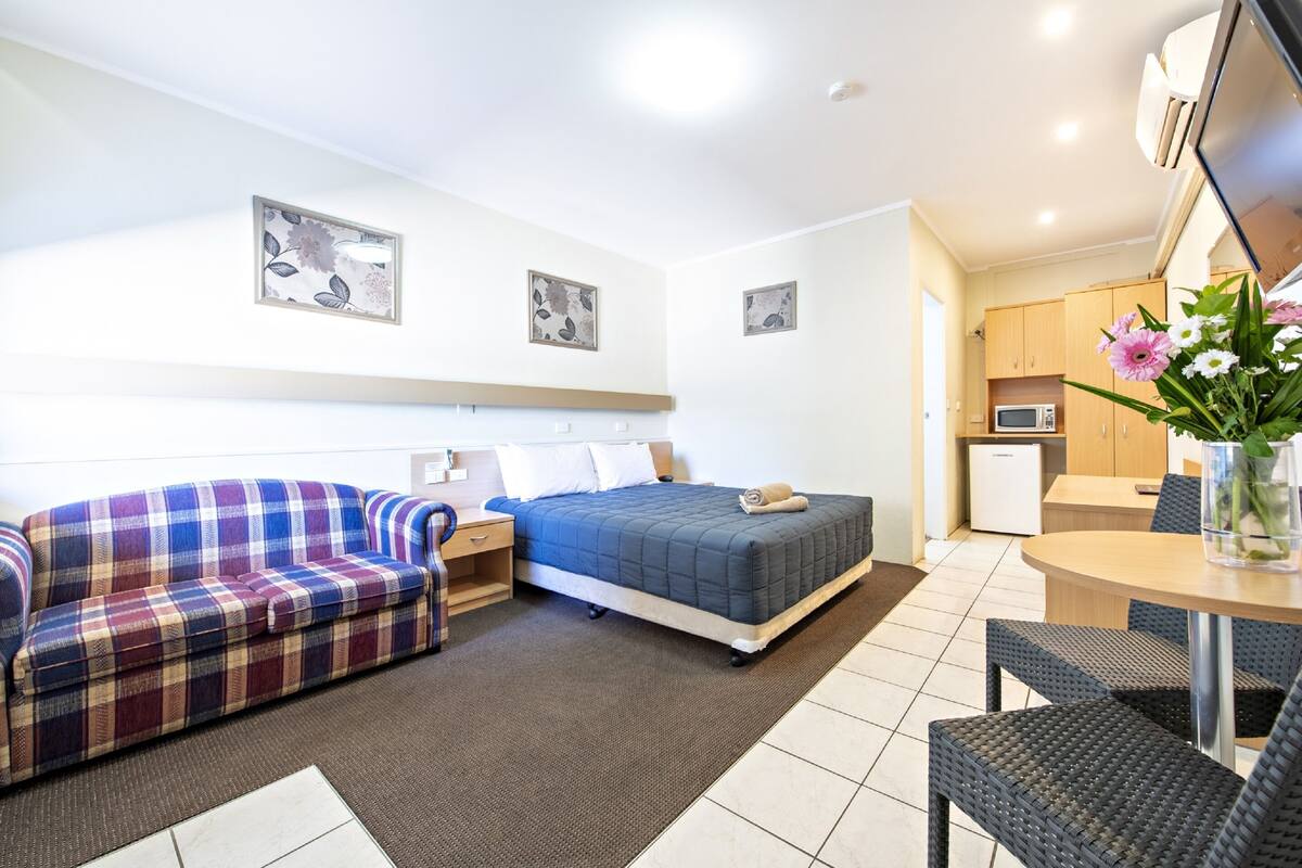 A modern hotel room with a bed, a sofa, a small kitchenette, and a dining area.