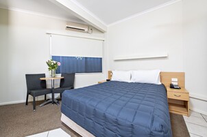 Habitación de hotel con una cama grande, un escritorio con una silla, una mesita con un florero y una ventana con persianas.