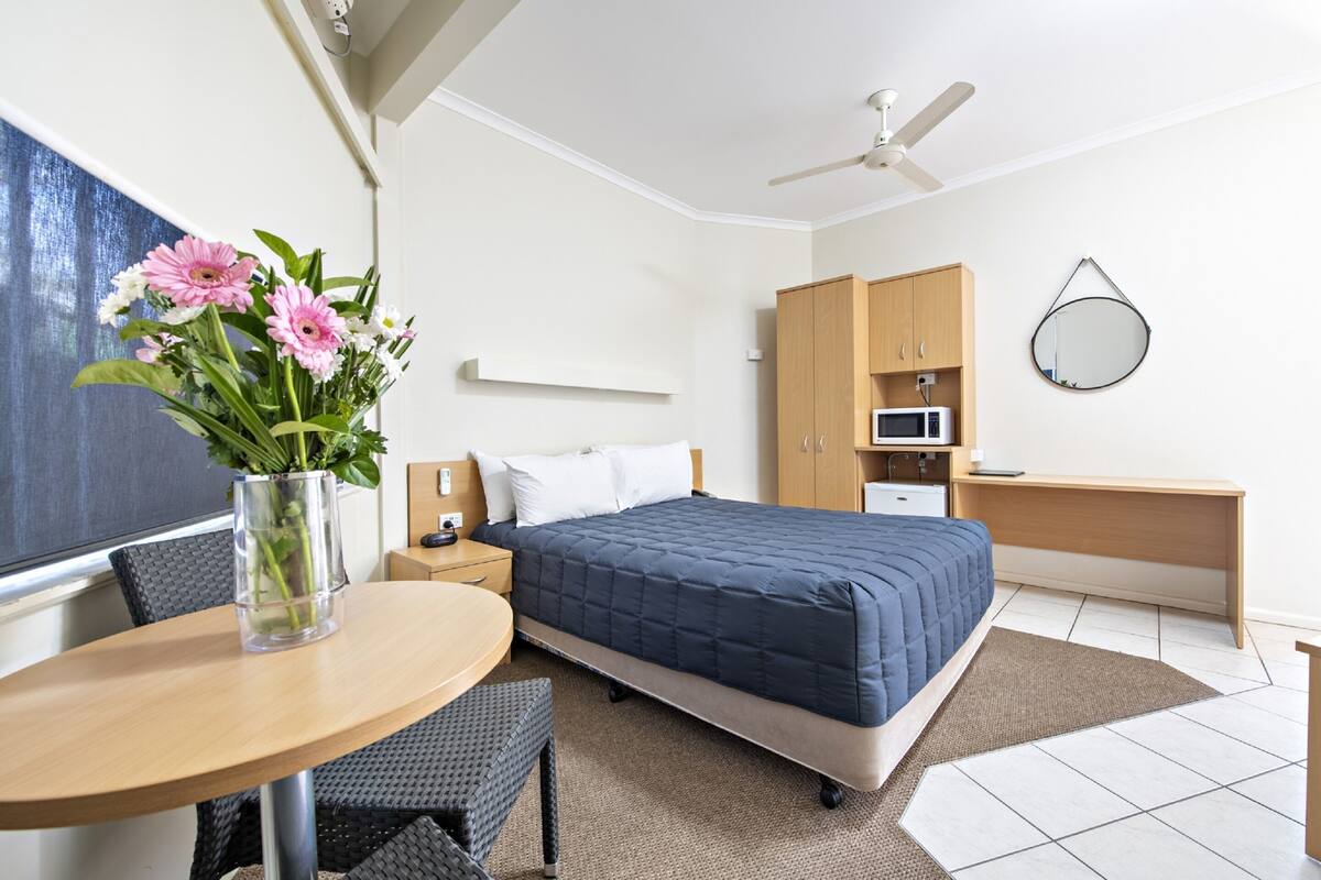 A hotel room with a bed, a desk, a chair, a small table, a vase with flowers, a ceiling fan, a microwave, and a mirror.