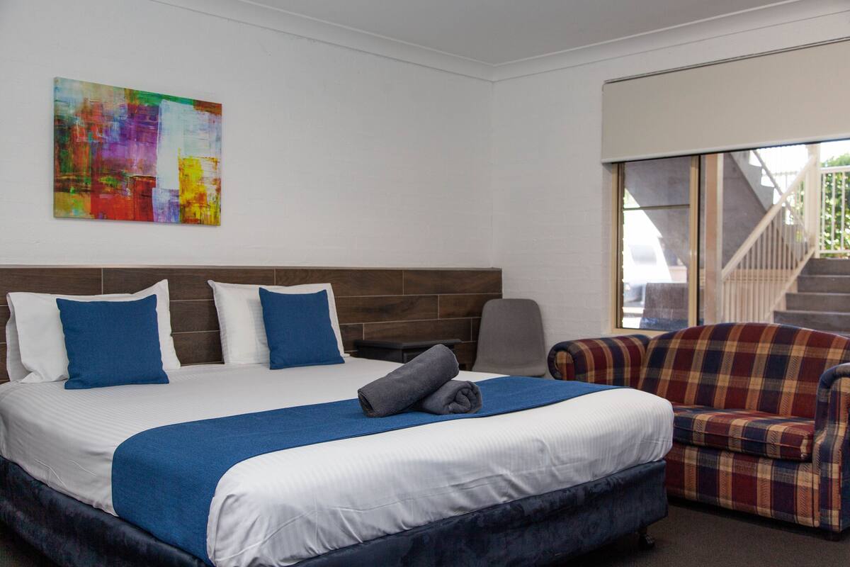 A hotel room with a large bed, a blue and white color scheme, a plaid armchair, and a colorful abstract painting on the wall.
