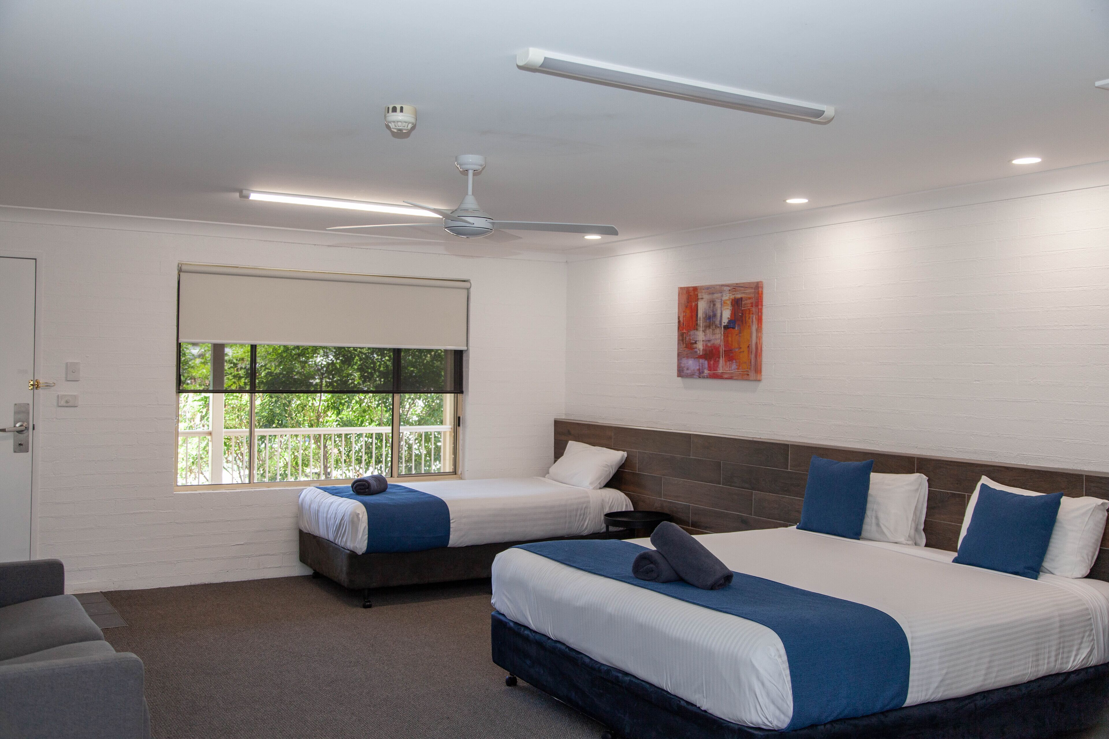 A hotel room with two beds, a ceiling fan, a window with a view, and a painting on the wall.