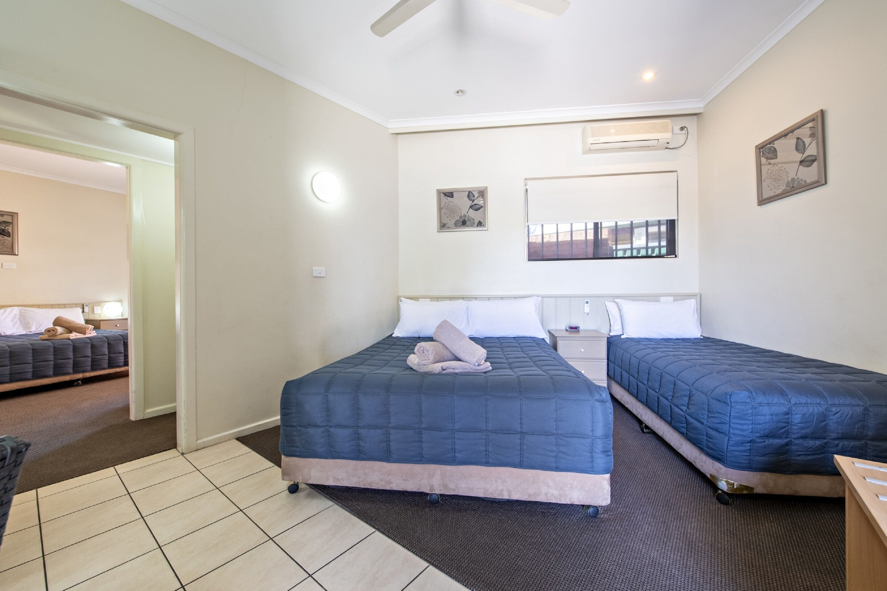 A hotel room with two beds, a ceiling fan, a window with bars, and artwork on the walls.