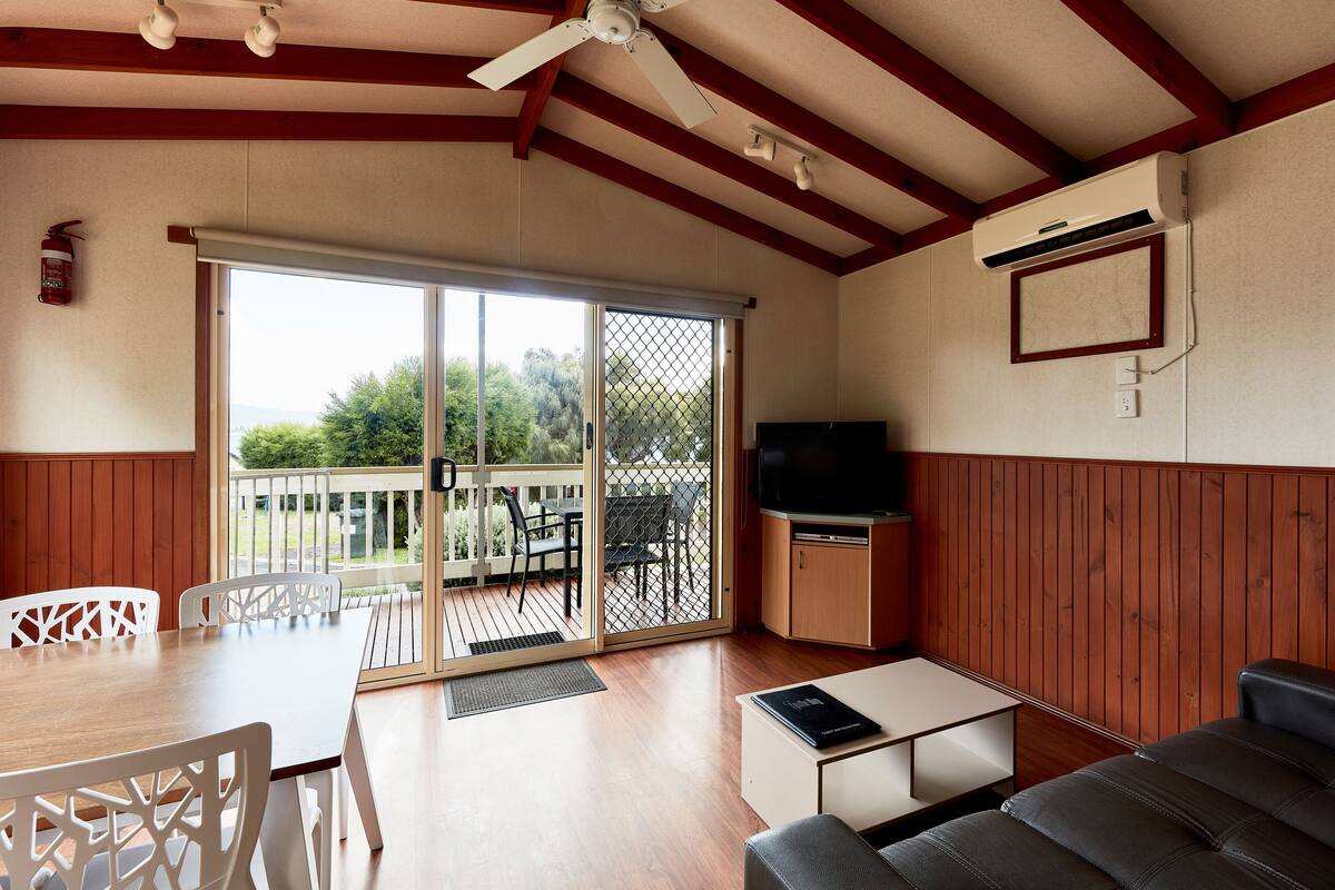 Una habitación con techo de madera, ventilador de techo, puerta de cristal corredera que da a un espacio exterior, televisión de pantalla plana, sofá y una mesa de comedor con sillas.