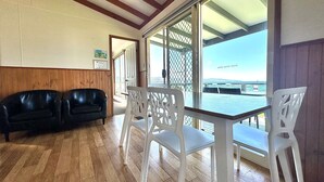 Una habitación con piso de madera, un sofá de cuero negro, sillas de plástico blanco y una mesa. Un ventanal ofrece vistas al océano y un puente.