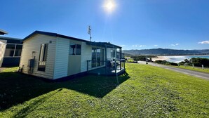 Una pequeña casa móvil de un piso con balcón, ubicada en un terreno cubierto de césped y con vista despejada a la playa y colinas al lejano.