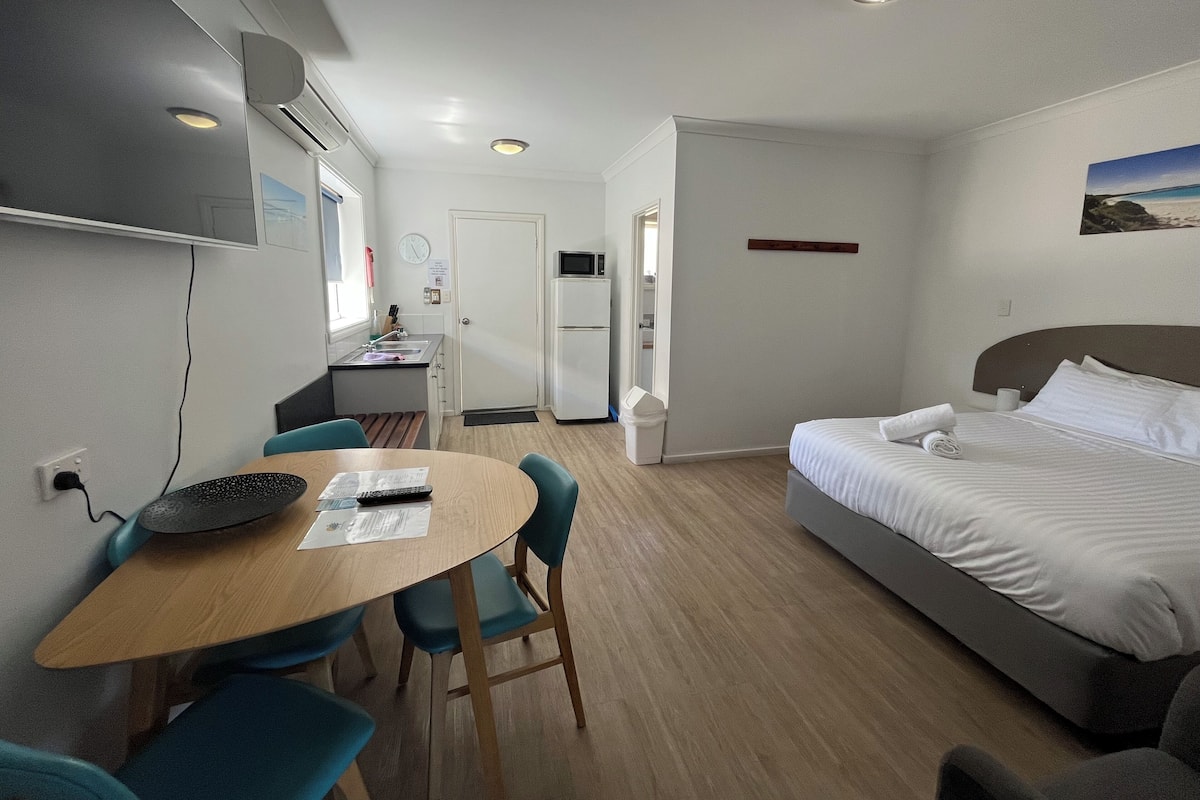 A hotel room with a bed, a desk with a chair, a small kitchenette, and a television mounted on the wall.