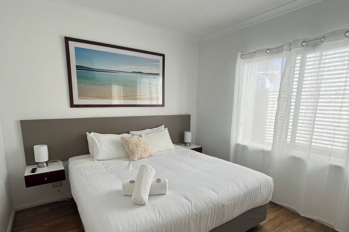 A neatly made bed with white linens, a bedside table with a lamp, and a framed picture of a beach on the wall.