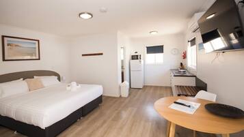 Un dormitorio con cama, una mesita con control remoto y una zona de cocina con refrigerador y microondas.