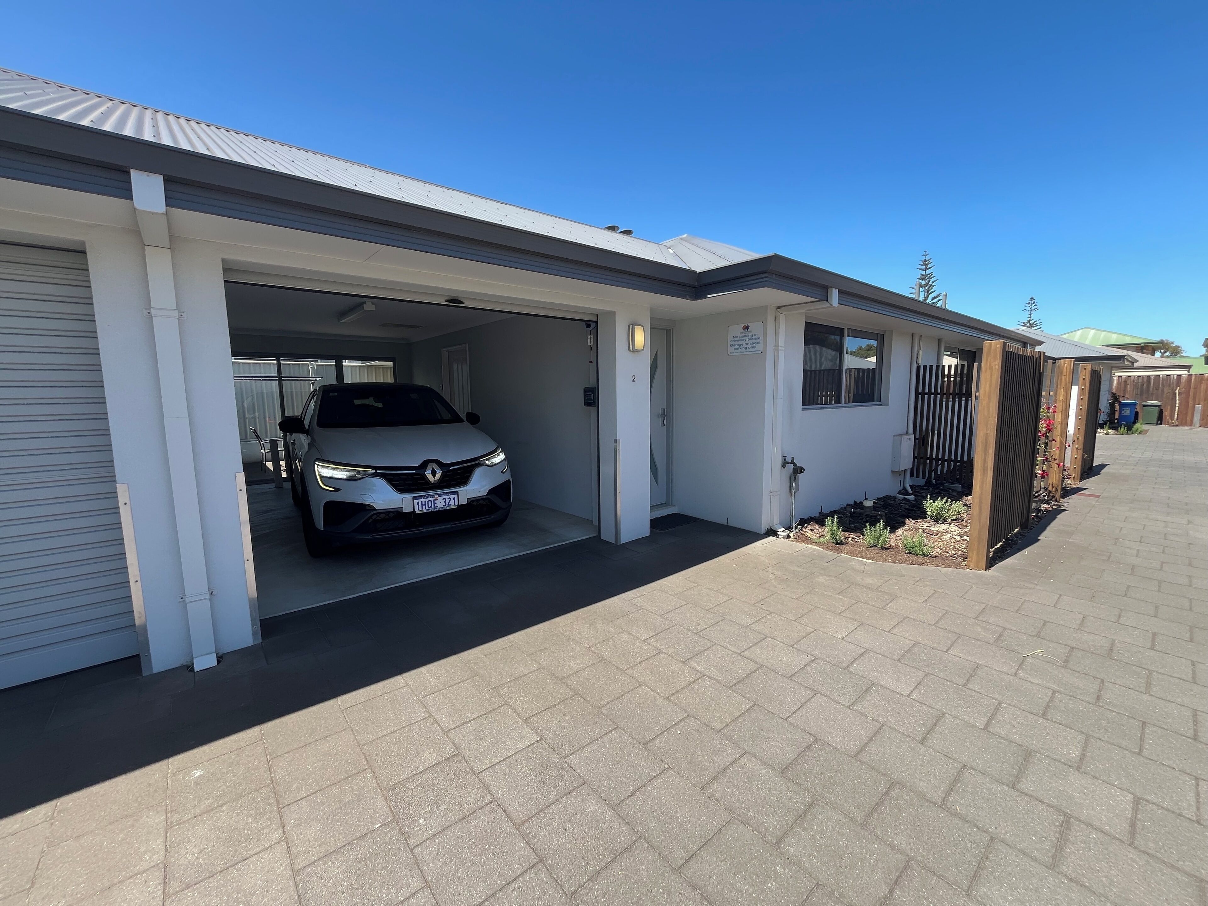 Un garage d’habitation avec une voiture garée à l’intérieur, une allée fermée par un portail et un ciel bleu dégagé.