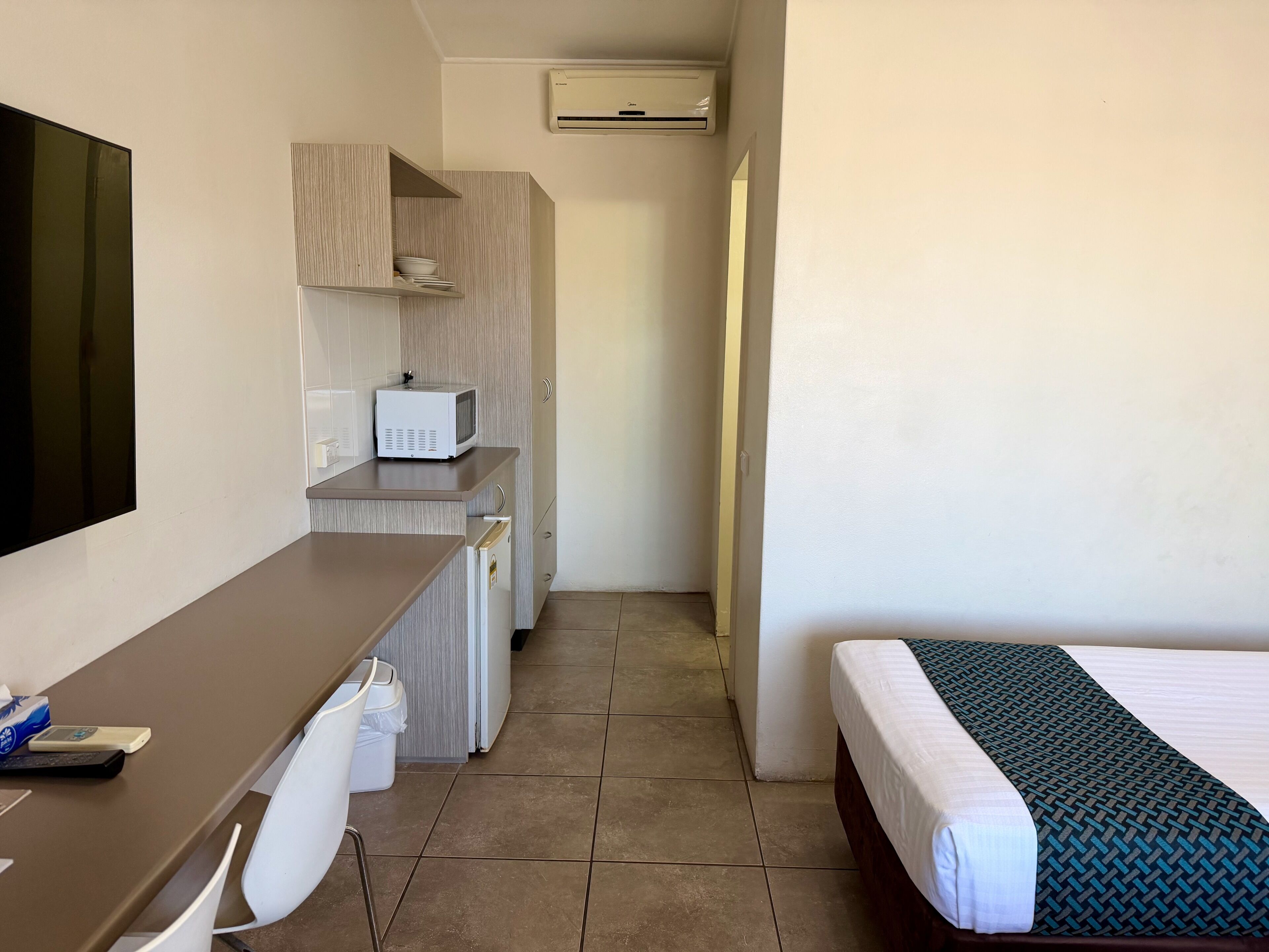 Habitación de hotel pequeña con cama, escritorio, televisor de pantalla plana, microondas y una kitchenette.