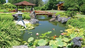 Um jardim tranquilo com um lago, uma ponte de madeira e um coreto.