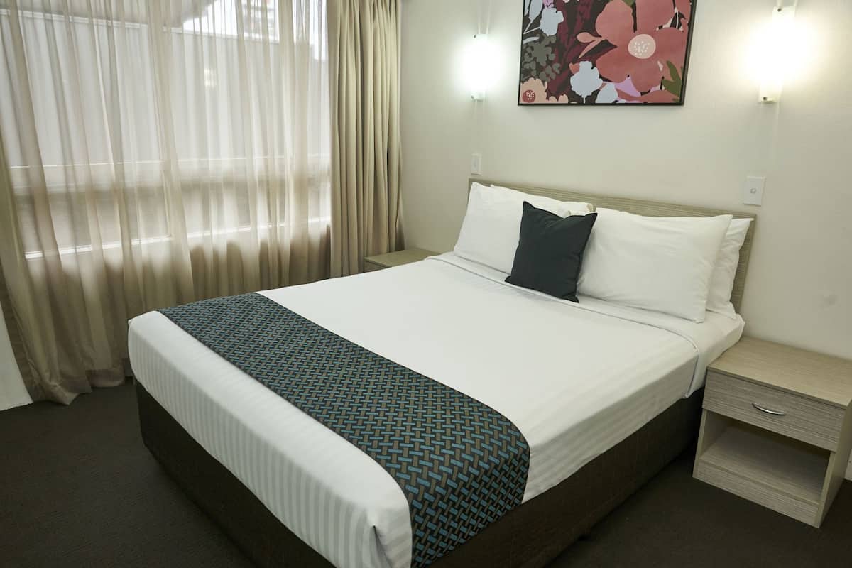 Habitación de hotel con cama, mesita de noche, cajón, un cuadro en la pared y cortinas traslúcidas.