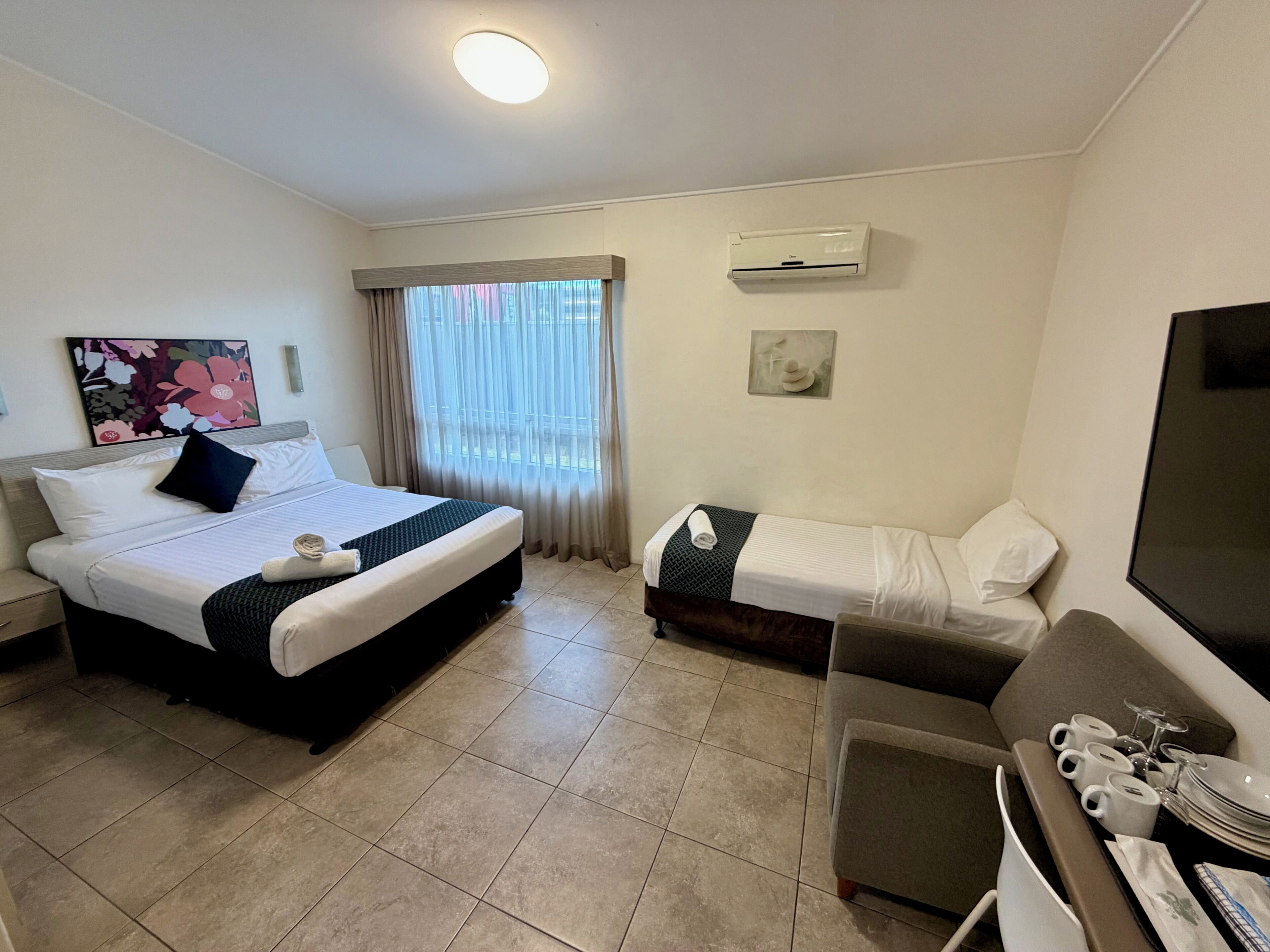 A hotel room with a bed, a sofa, a TV, a window with curtains, and an air conditioning unit.