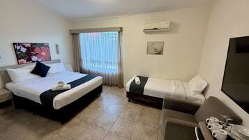 A hotel room with a bed, a sofa, a TV, a window with curtains, and an air conditioning unit.