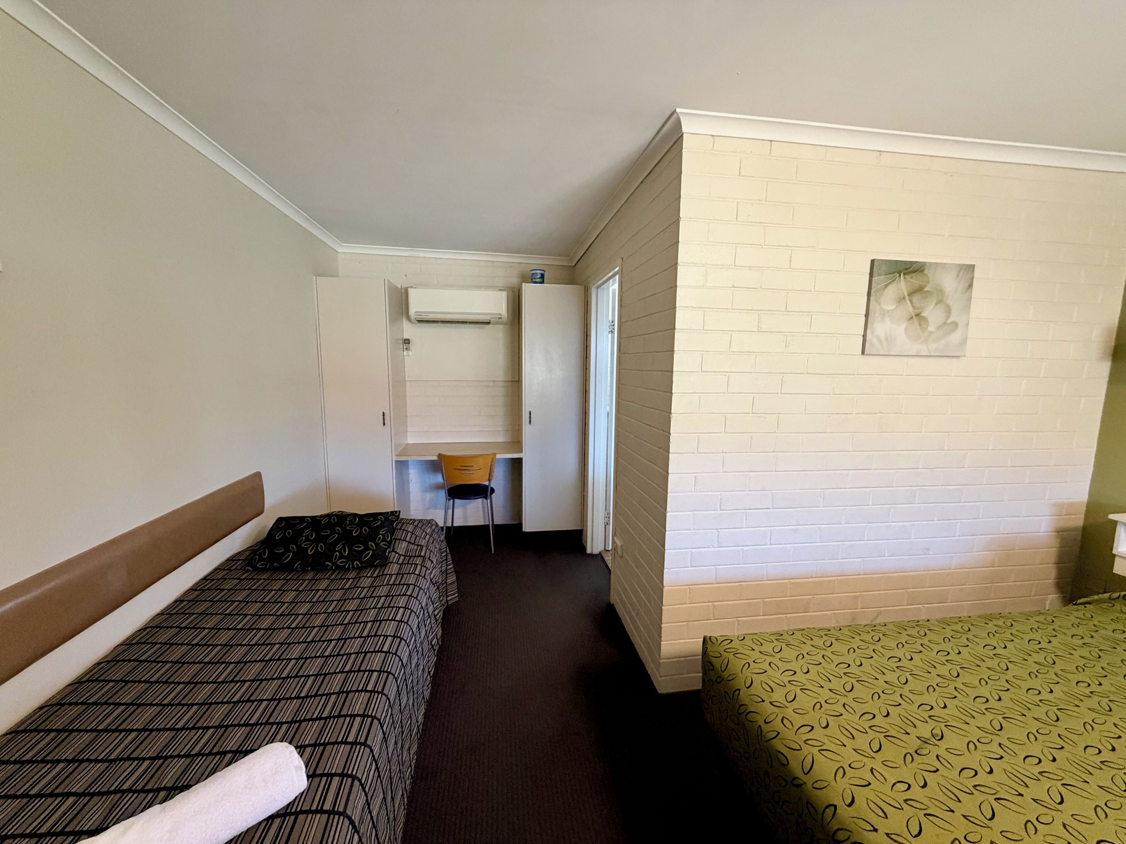 A compact hotel room with two beds, a small desk, a chair, and a white brick wall.