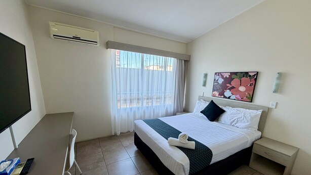 Quarto de hotel com cama, escrivaninha, cadeira, televisão, ar condicionado, janela com cortinas, mesa de cabeceira, gaveta e um quadro na parede.