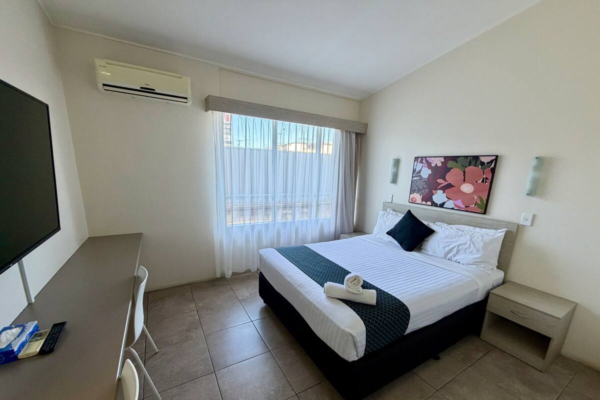 Habitación de hotel con cama, escritorio, silla, televisor, aire acondicionado, ventana con cortinas, mesita de noche, cajón y un cuadro en la pared.