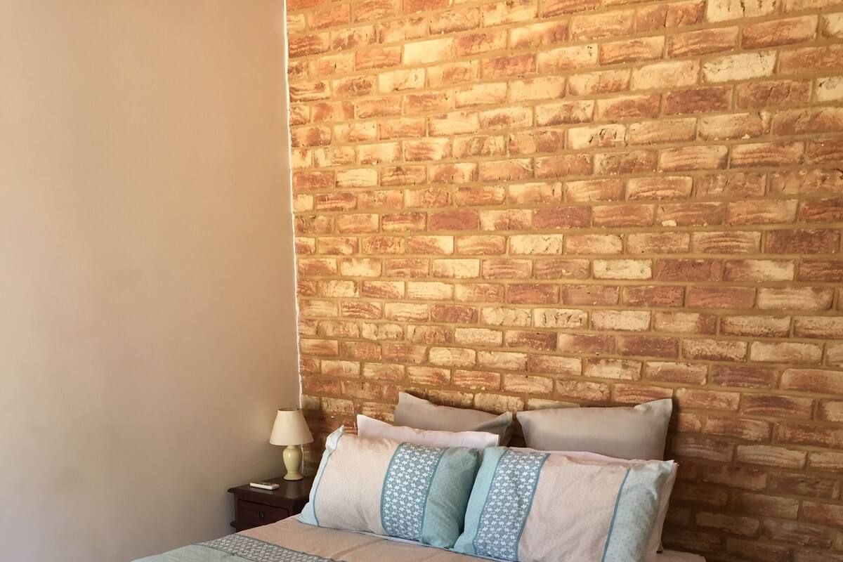 Een bed met een gebedekt sprei en kussens, een nachtkastje met een lamp, en een bakstenen muur achter het bed.