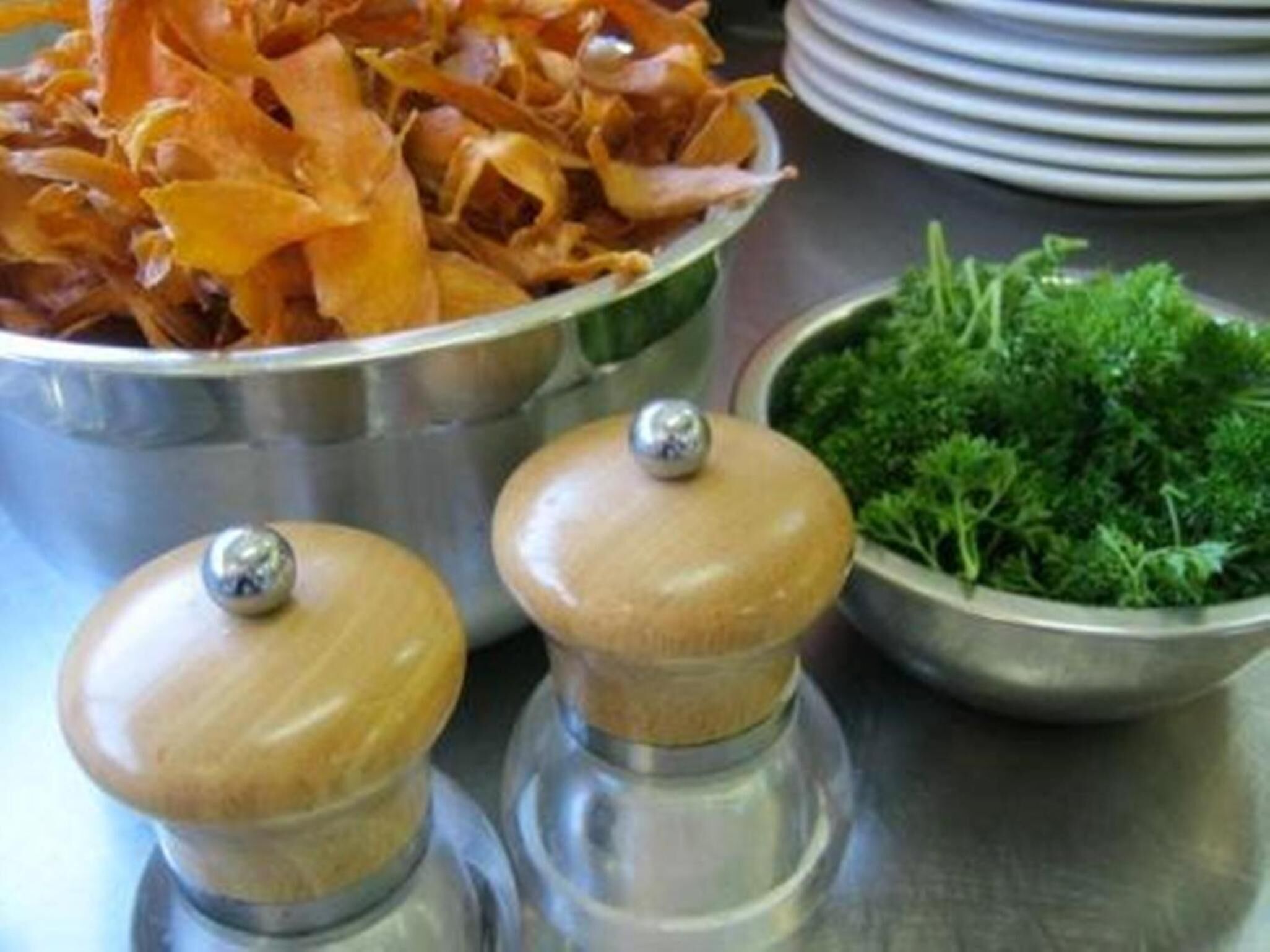 A stainless steel bowl filled with dried orange slices, a bowl of green herbs, and two wooden salt and pepper shakers on a metallic surface.