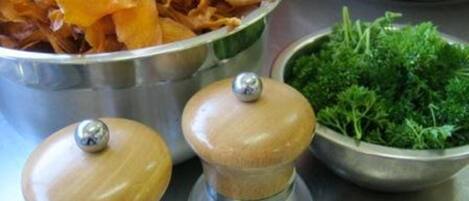 Un tazón de acero inoxidable lleno de rodajas de naranja deshidratada, un tazón de hierbas verdes y dos saleros y pimenteros de madera sobre una superficie metálica.