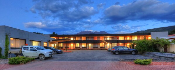 Un hotel con estacionamiento, un edificio con múltiples ventanas y una montaña al fondo.