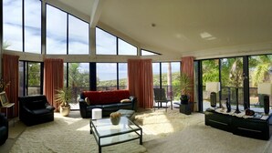 A spacious living room with large windows, a black leather sofa, a glass coffee table, and a view of palm trees and hills. - Seaview Bed and Breakfast (Bonny Hills)