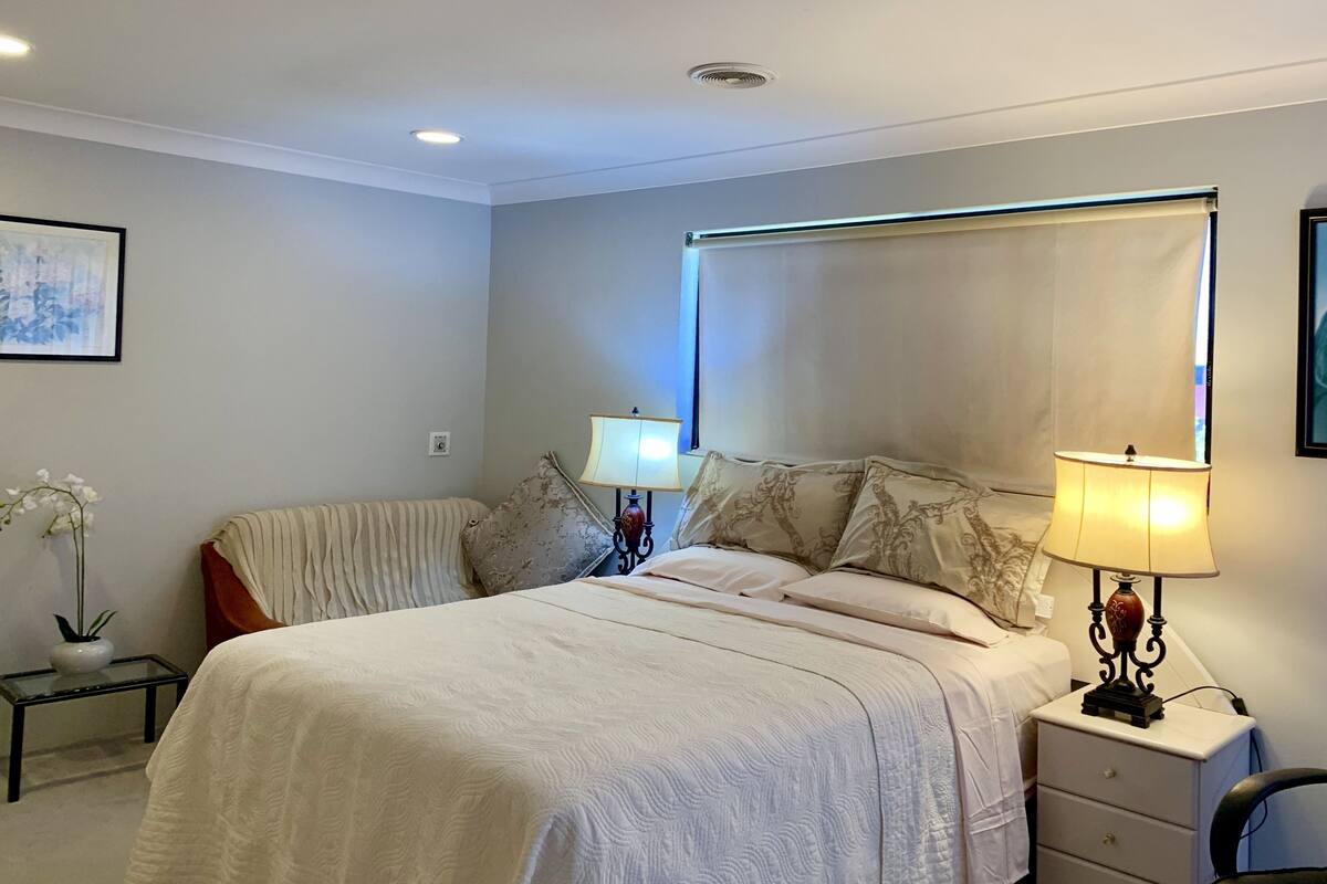 A bedroom with a large bed, two bedside tables, a nightstand, a lamp, a wall-mounted shelf, and a framed picture.