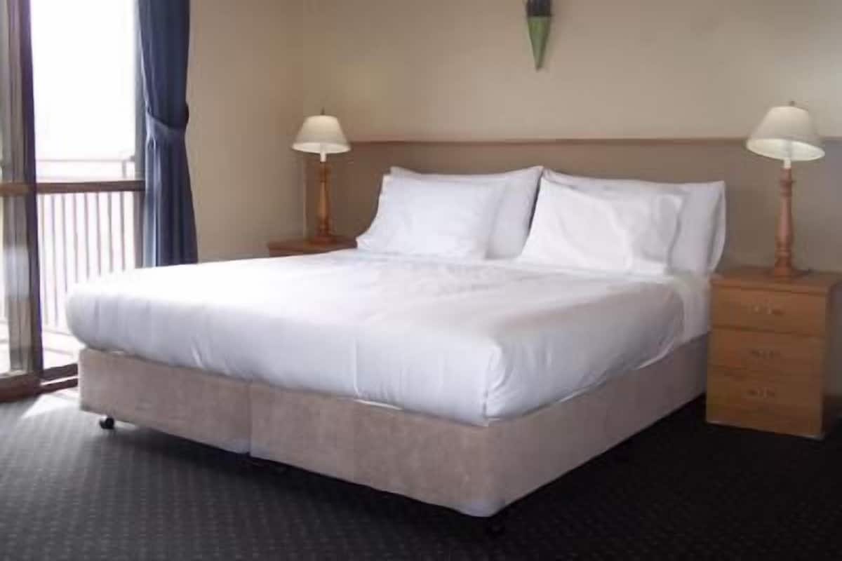 A hotel room with a large bed, two bedside tables with lamps, a window with curtains, and a wall-mounted light fixture.