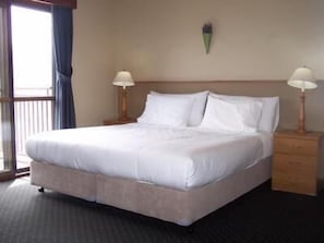A hotel room with a large bed, two bedside tables with lamps, a window with curtains, and a wall-mounted light fixture.
