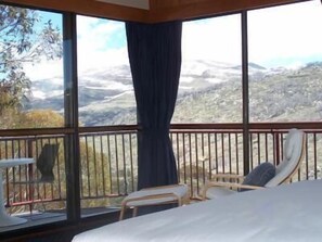 Quarto com cama, cadeira e mesa. Grande janela com vista para paisagem montanhosa.