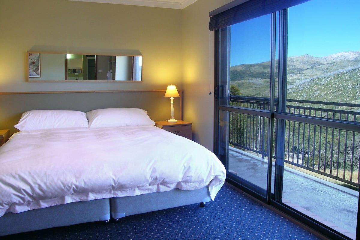 A hotel room with a large bed, a nightstand, a lamp, a mirror, and a balcony with a mountain view.