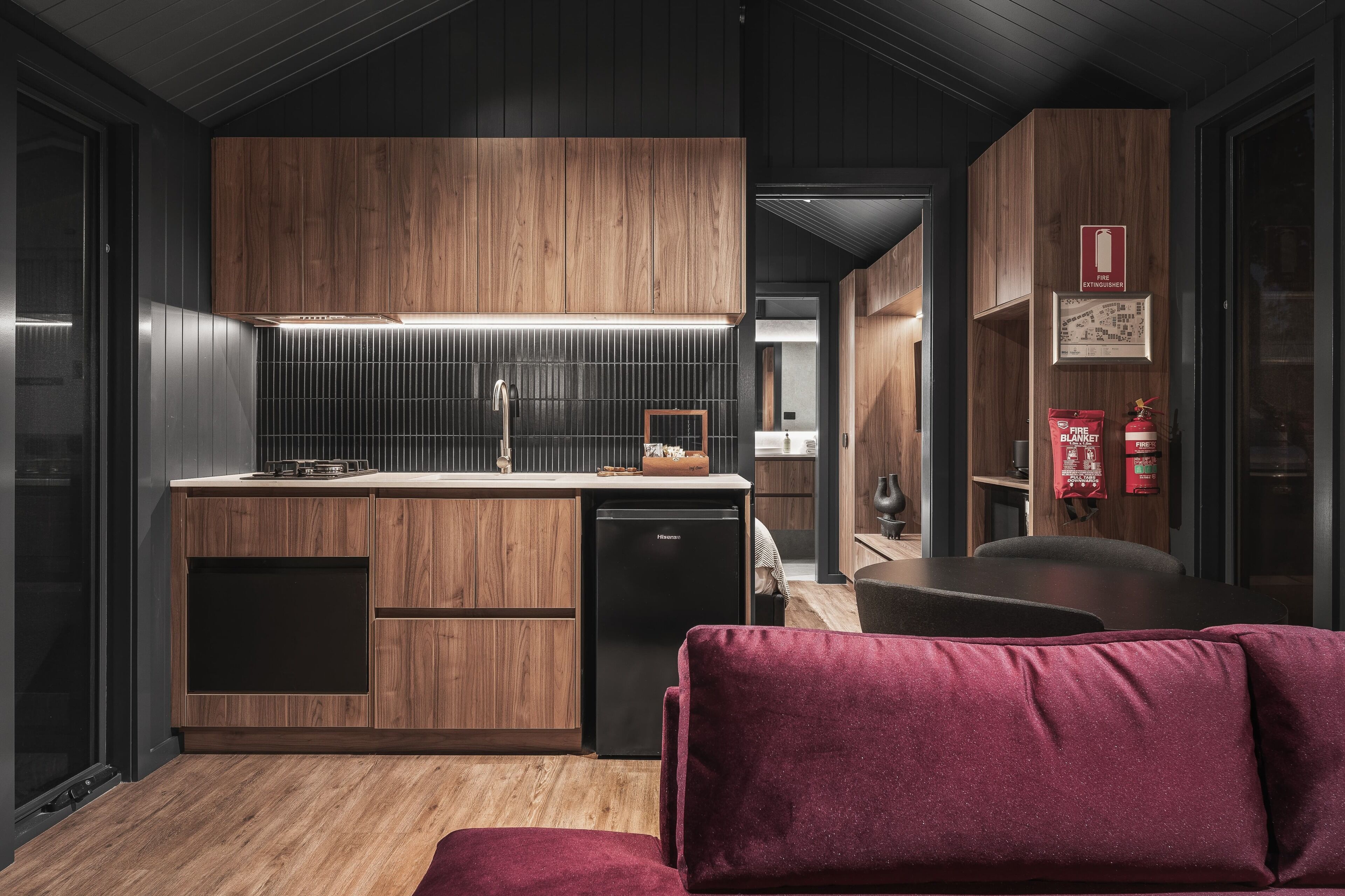 A modern kitchen with wooden cabinets, a black refrigerator, and a dining area with a red sofa.