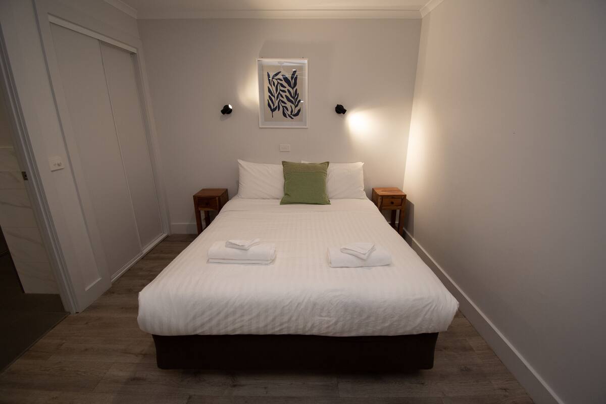 A neatly made bed with white linens and two pillows, a green accent pillow, and two folded towels on the bed.
