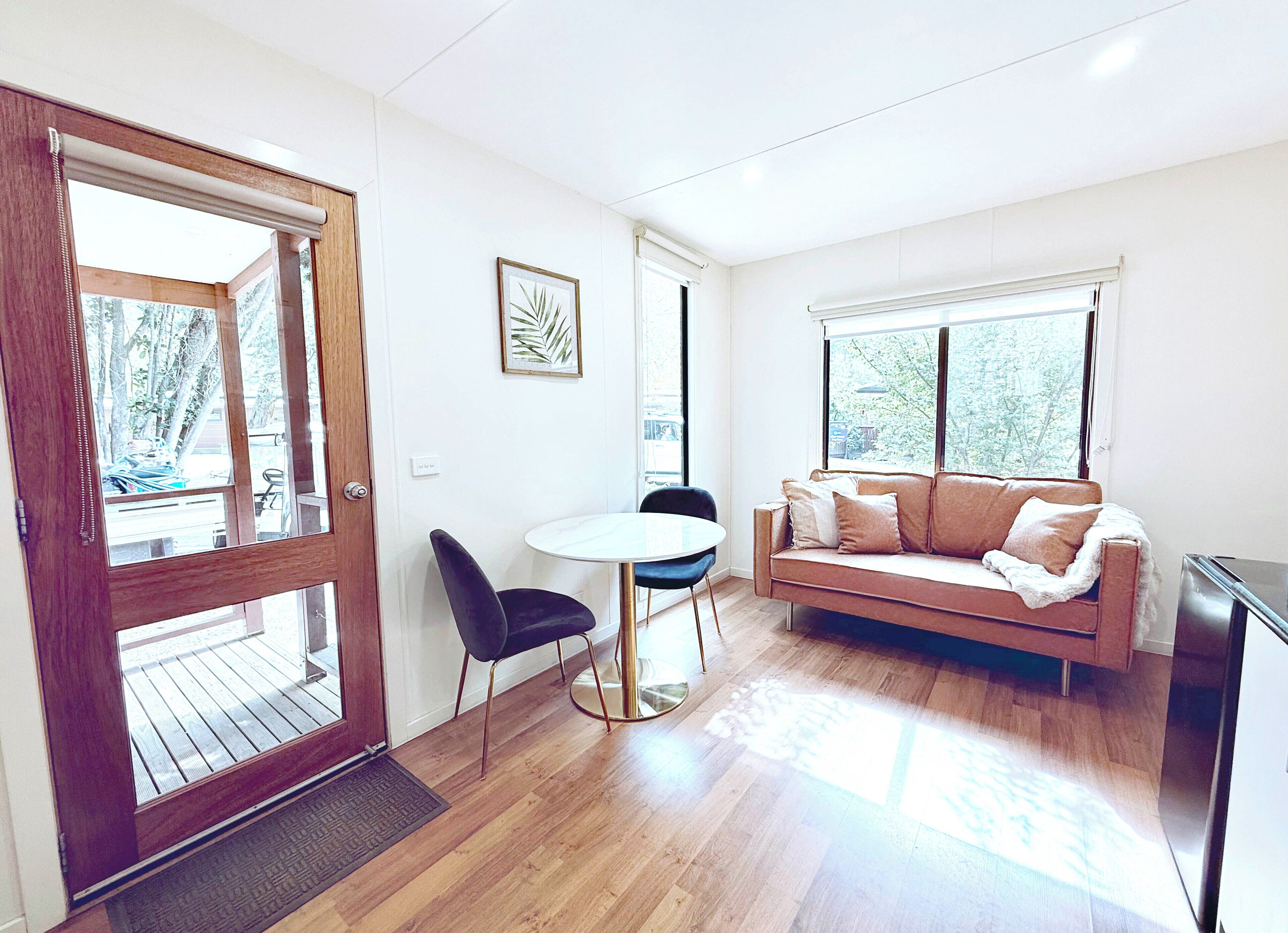 A room with a wooden floor, a white round table with two chairs, a beige sofa with a blanket, and a large window with a view of trees.