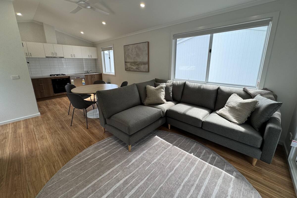 A modern living room with a grey sectional sofa, a dining table with chairs, a kitchen area with white cabinets, and a large window providing natural light.