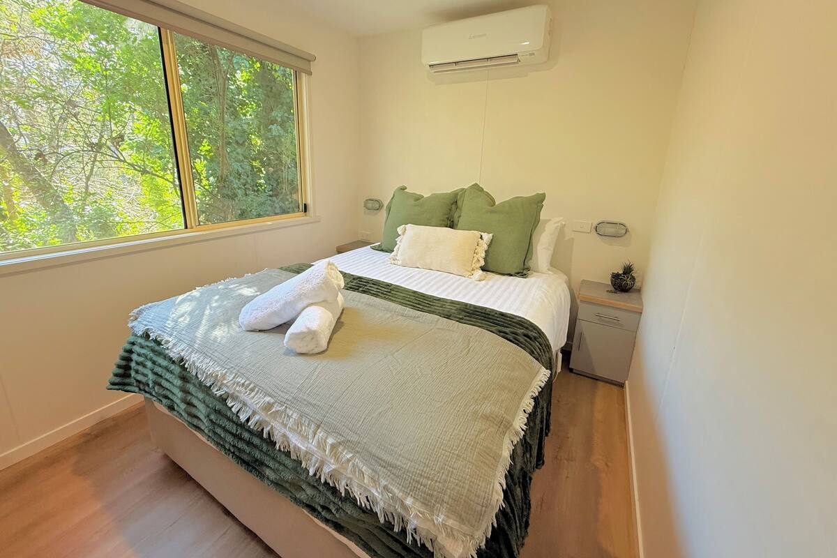 A neatly made bed with a green and white blanket, a white pillow, and a green pillow. There is a small bedside table with a plant and a lamp. A window with a view of trees is visible.