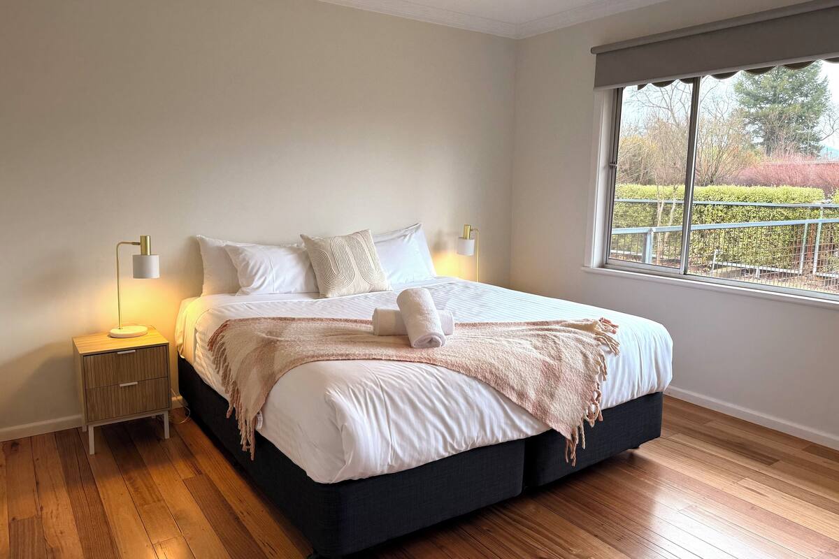 A bedroom with a large bed, wooden flooring, and a window with a view of greenery.