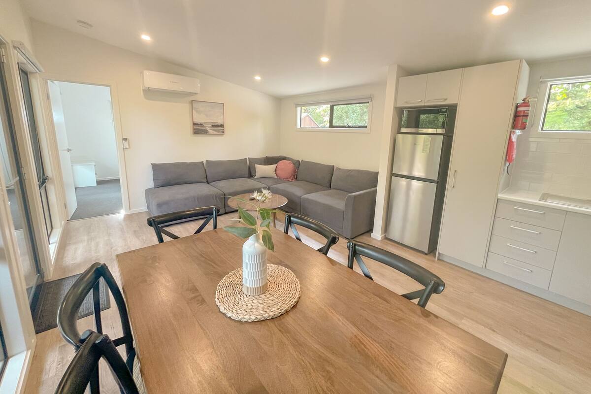 A modern living space with a dining area, grey sofa, kitchen, and a view of the outdoors.