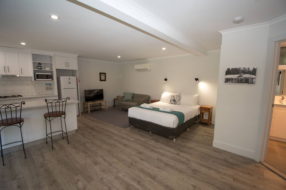 A compact bedroom with a bed, a small sofa, a TV, and a kitchenette with a microwave and refrigerator.