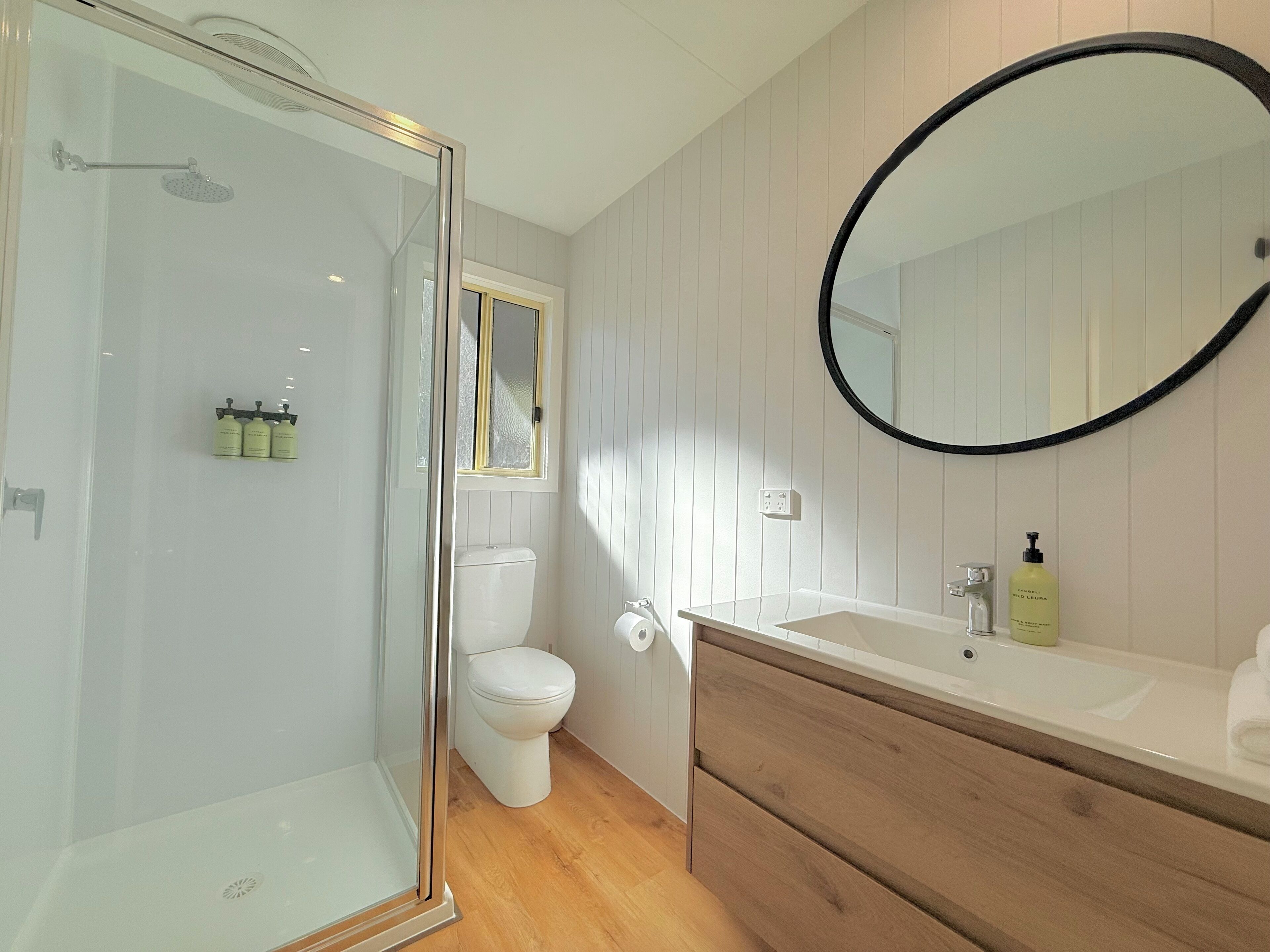 A bathroom with a glass-enclosed shower, a toilet, a vanity with a mirror, and toiletries on the counter.