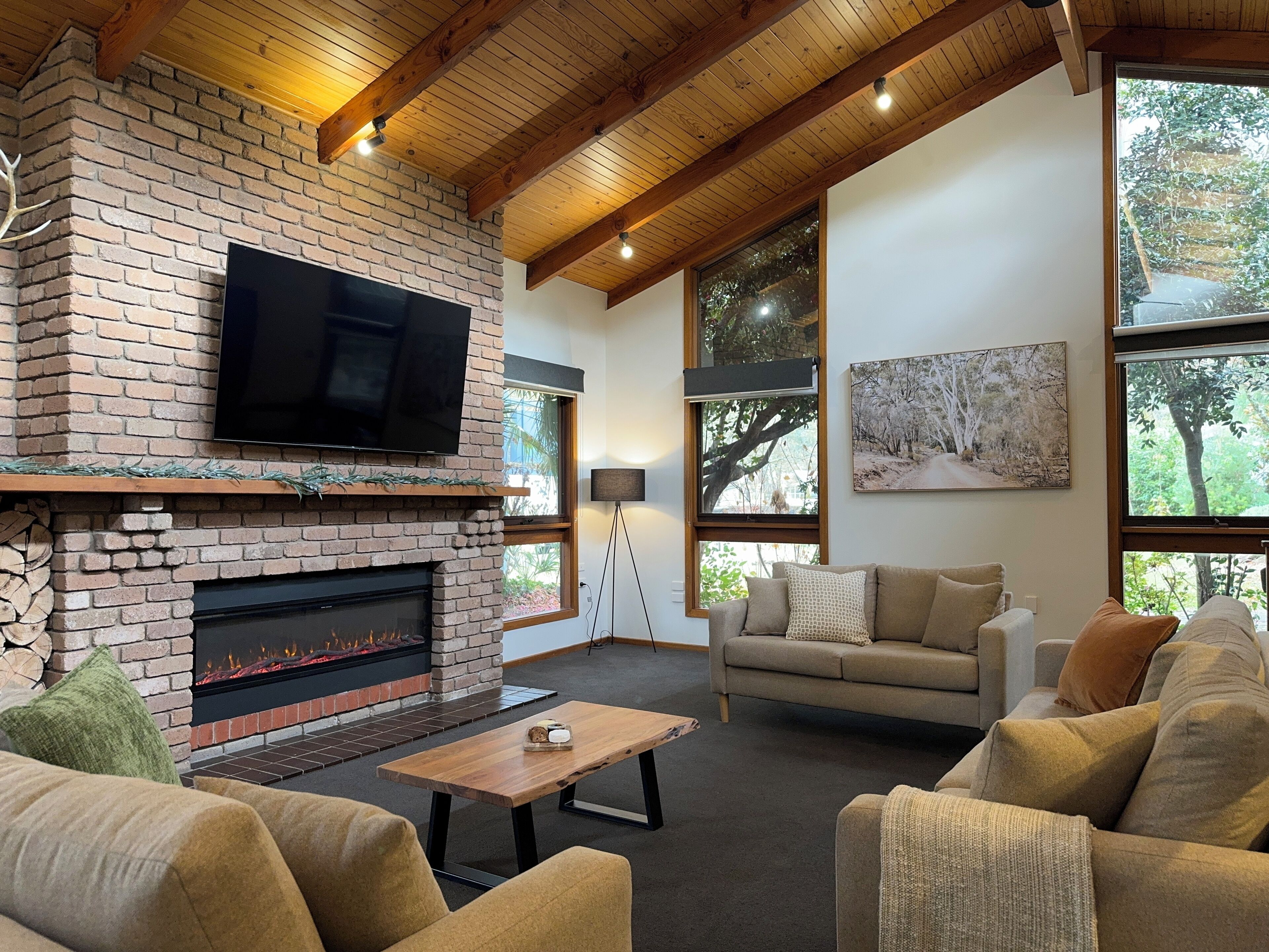 A cozy living room with a brick fireplace, a flat-screen TV, a sofa, and large windows offering views of trees.