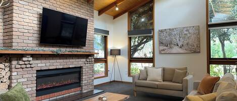 A cozy living room with a brick fireplace, a flat-screen TV, a sofa, and large windows offering views of trees.