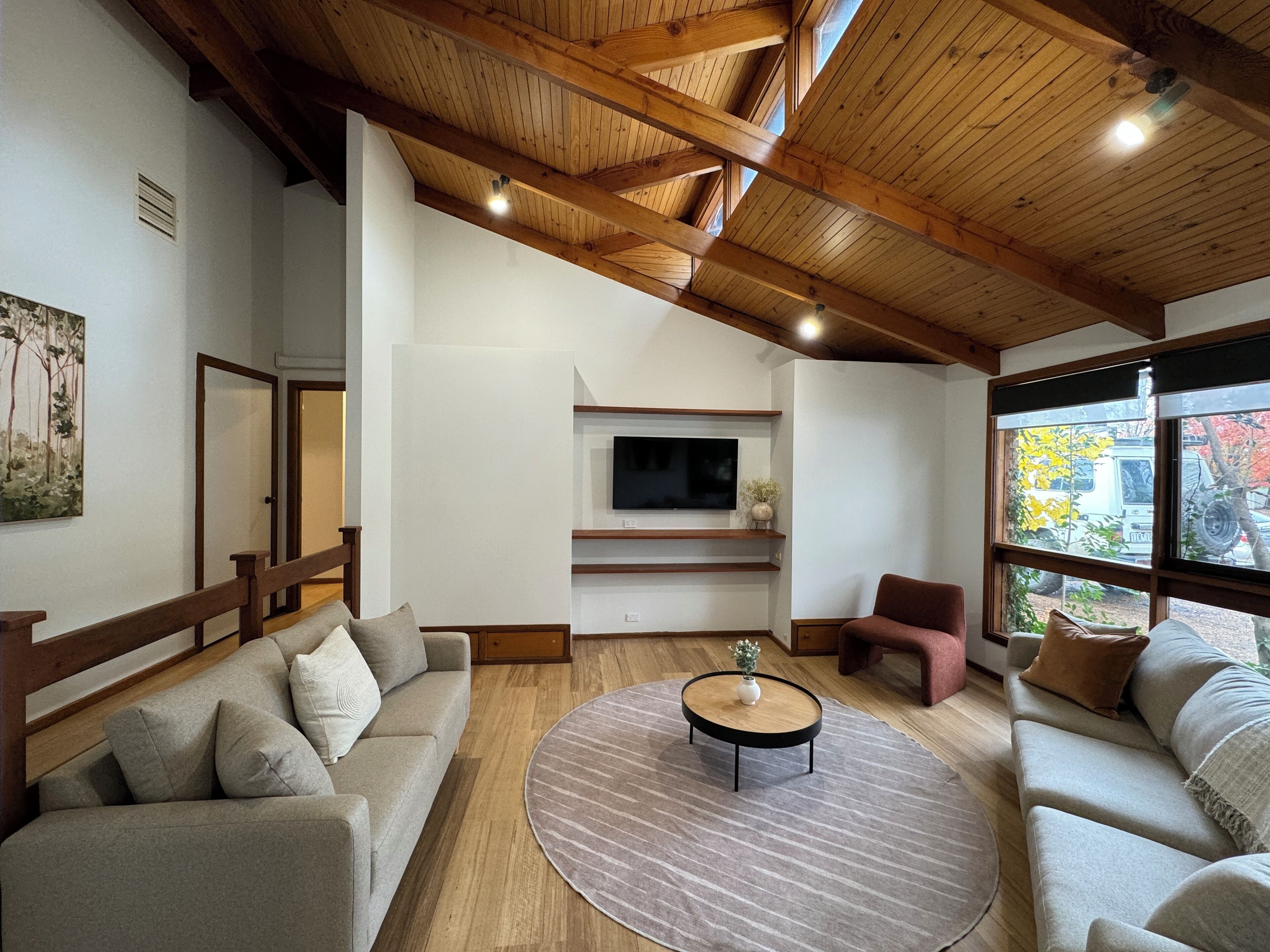 Un vaste salon doté d’un plafond en bois, d’un téléviseur à écran plat fixé au mur, d’une table basse ronde et d’une grande fenêtre offrant une vue sur l’extérieur.